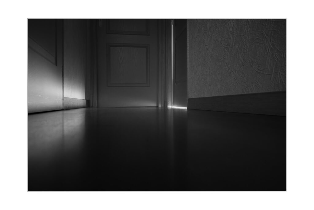 Photo noir et blanc en contre-plongée d'un couloir avec des portes fermées ; l'une des portes est légèrement entrouverte, laissant pénétrer un éclat de lumière sur le sol, créant des ombres et des contrastes spectaculaires.