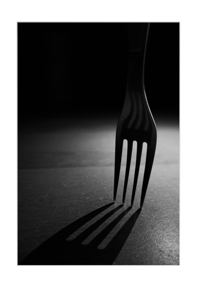 Photo en noir et blanc d'une fourchette posée à la verticale sur une surface plane, projetant une longue ombre dramatique au premier plan. L'éclairage crée une atmosphère sombre et contrastée.