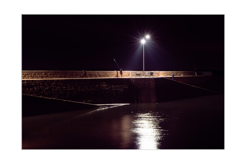 Une personne pêche sur une jetée en briques, la nuit, éclairée par un seul lampadaire dont la lumière se reflète sur l'eau calme en contrebas. L'environnement est sombre, ce qui souligne l'atmosphère paisible et tranquille.