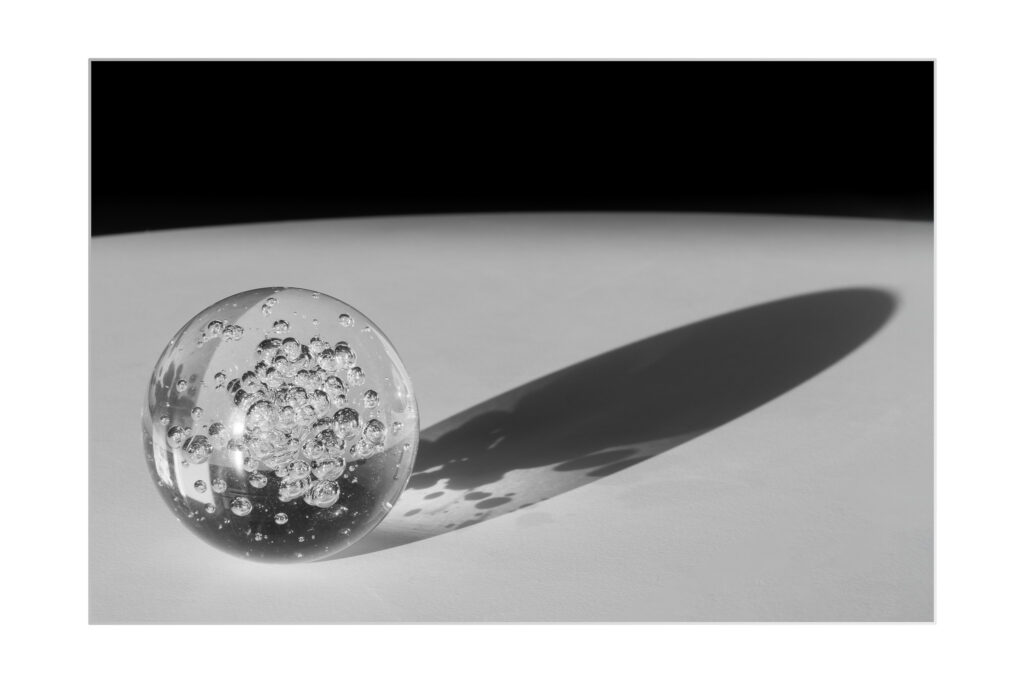Une sphère en verre transparent avec des bulles d'air à l'intérieur est posée sur une surface blanche lisse, projetant une longue ombre. L'arrière-plan passe du blanc au noir, créant un contraste saisissant.