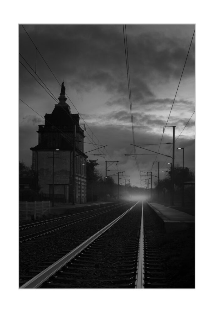 Gare Sainte-Anne à Pluneret. Photo en noir et blanc de voies ferrées vides au crépuscule, avec des fils électriques aériens et un vieux bâtiment à gauche. La voie ferrée s'estompe dans le lointain sous un ciel dramatique et nuageux.
