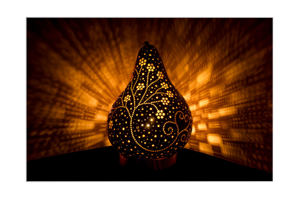 Une lampe décorative en forme de calebasse aux motifs complexes s'allume chaudement, projetant des motifs lumineux détaillés et des ombres sur le mur environnant dans une pièce sombre.
