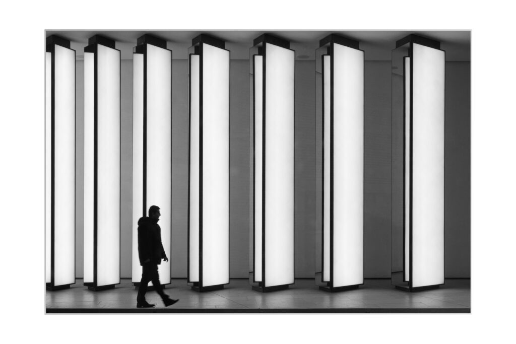 Une personne passe devant de grands panneaux rectangulaires verticaux éclairés dans un décor moderne et monochrome. La scène est minimaliste, la silhouette de la personne se détachant sur les colonnes lumineuses.