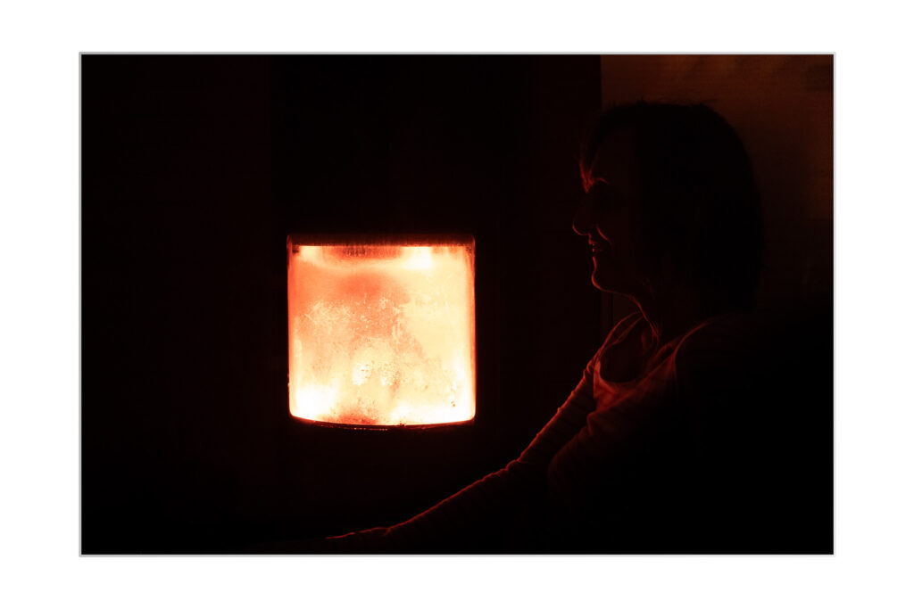 Une personne est assise dans une pièce sombre, éclairée par la lueur chaude et orangée d'une cheminée ou d'un poêle. Le silhoutte de son profil est visible à contre-jour.