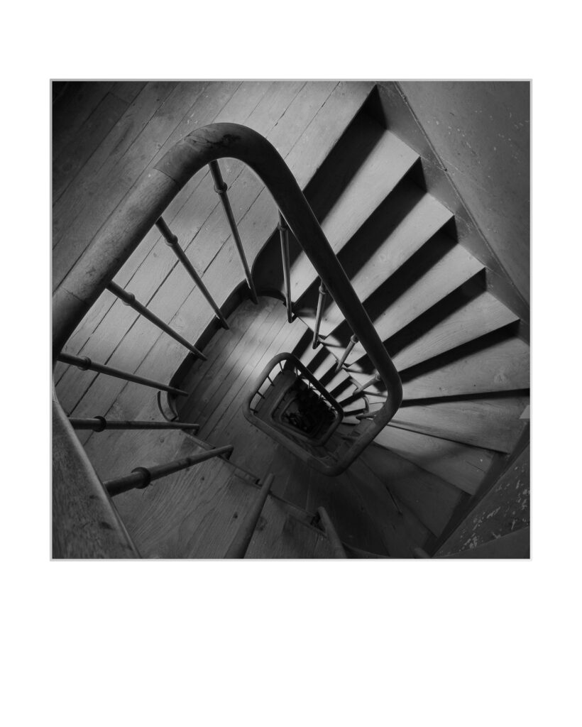 Photo en noir et blanc d'un escalier en colimaçon vu de dessus, montrant des marches en bois et une main courante en métal avec des fuseaux verticaux, créant un motif géométrique, presque hypnotique, au fur et à mesure que l'escalier descend.