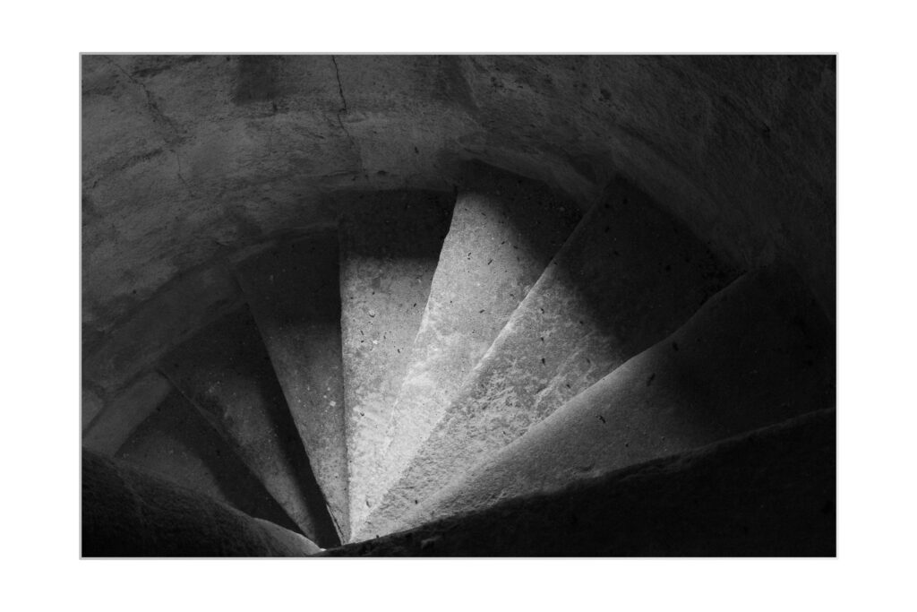 Photo en noir et blanc d'un escalier de pierre en colimaçon vu de haut, avec un éclairage dramatique qui projette des ombres et met en valeur les marches texturées et le mur incurvé.