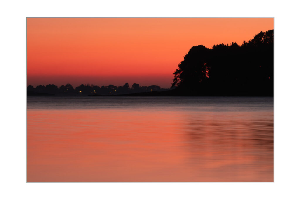 Une étendue d'eau calme reflète les teintes orange et rouge vives d'un ciel de coucher de soleil, avec des silhouettes sombres d'arbres et des lumières lointaines visibles à l'horizon.