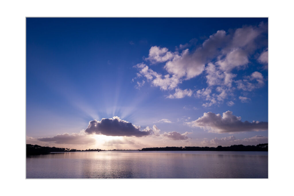 Scène de lac sereine au lever ou au coucher du soleil, avec des rayons de soleil traversant des nuages épars dans un ciel bleu vibrant, se reflétant sur l'eau calme en contrebas. Des arbres bordent l'horizon lointain.