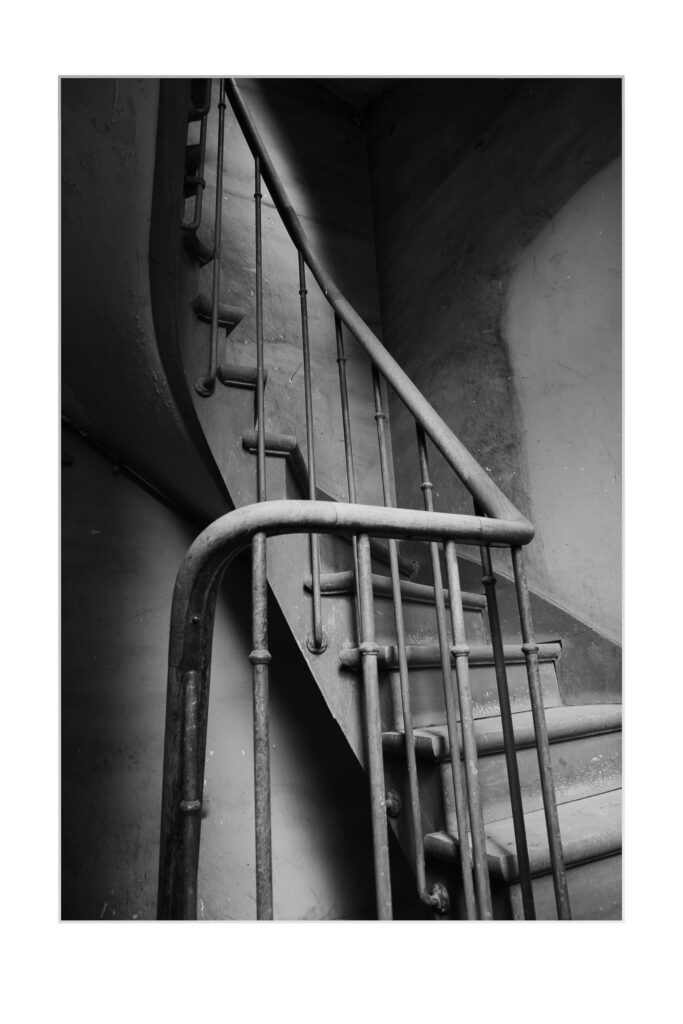 Photo en noir et blanc d'un escalier en colimaçon avec des rampes en métal dans un vieux bâtiment faiblement éclairé. L'ombre et la lumière mettent en valeur les courbes et la texture usée des marches et des murs.