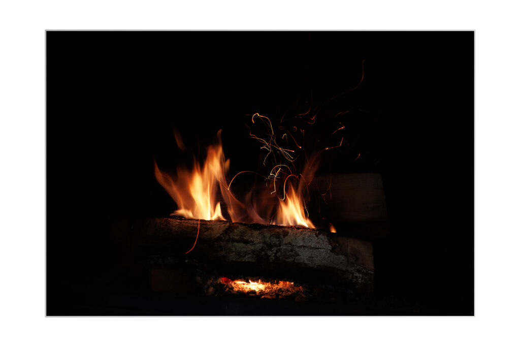 Gros plan d'une bûche en feu dans un environnement sombre, avec des flammes orange vif et des étincelles visibles se détachant sur un fond noir.