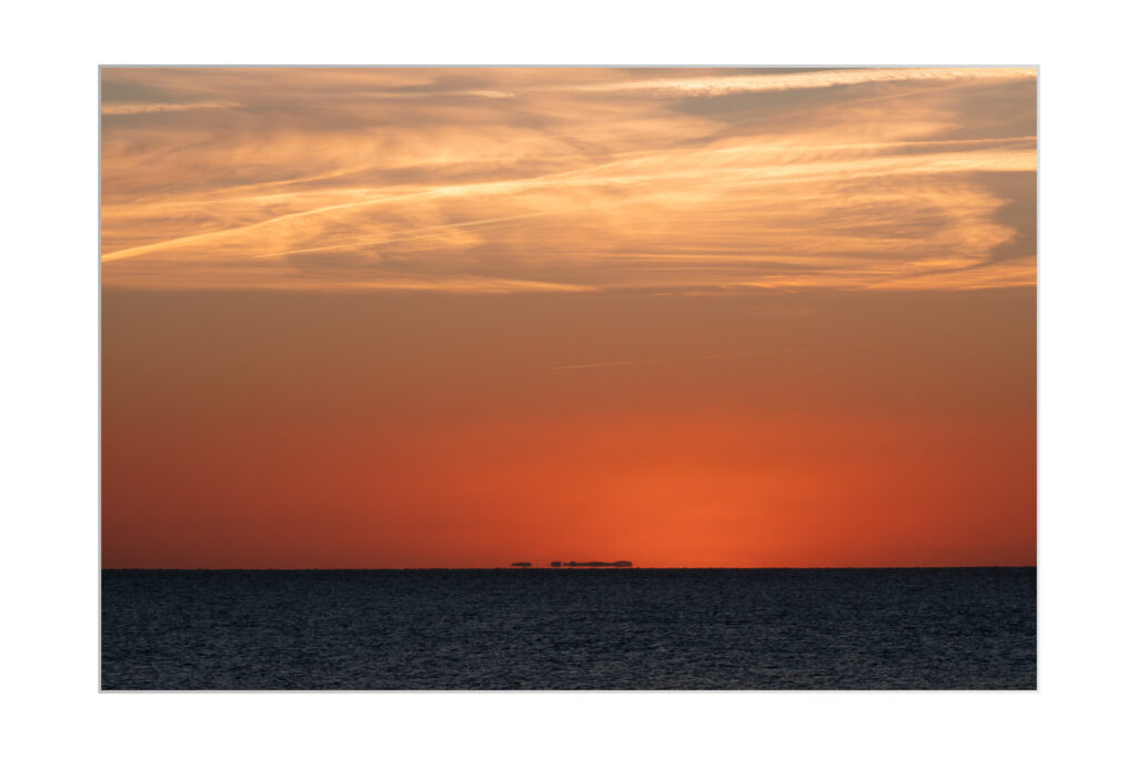 Un coucher de soleil éclatant sur l'océan, avec des nuages orange et jaunes qui traversent le ciel. Une faible silhouette de structures lointaines apparaît à l'horizon, au-dessus de l'eau sombre.