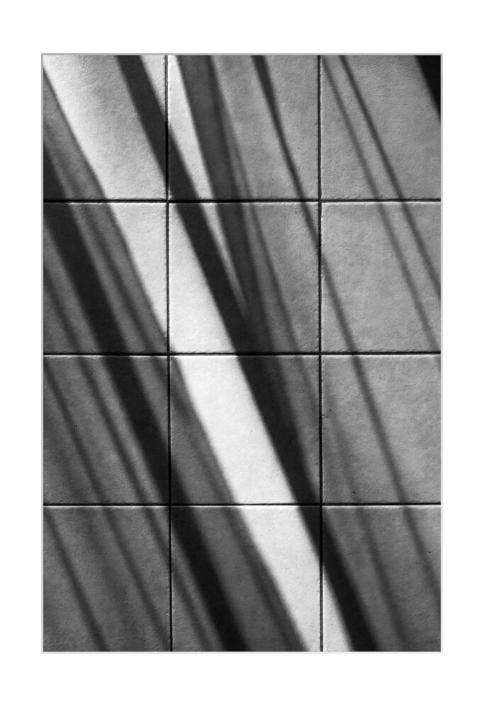 Photo en noir et blanc d'une surface carrelée, avec de fortes ombres diagonales projetées sur les carreaux, créant un motif dynamique de lumière et d'obscurité.