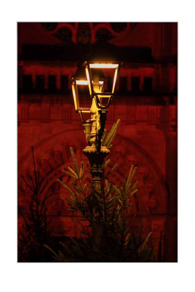 Un lampadaire vintage brille chaleureusement la nuit, entouré de branches de pin vertes, avec un bâtiment orné à l'arrière-plan éclairé par une lumière rouge.