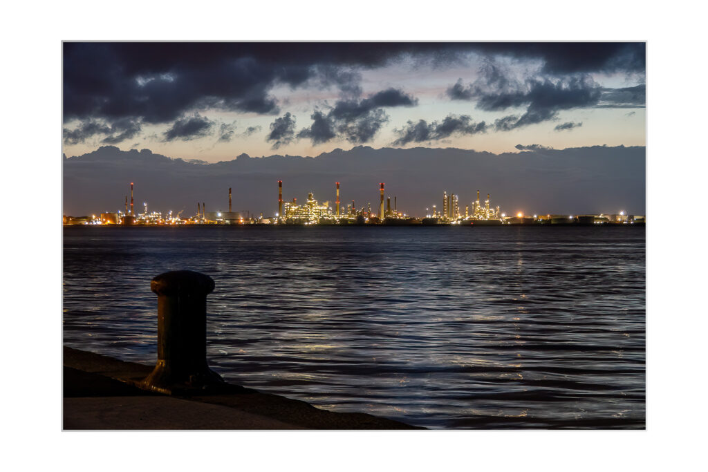 Un front de mer au crépuscule avec des nuages sombres au-dessus, une eau calme au premier plan et une raffinerie industrielle très éclairée avec de hautes cheminées au loin. Une grande borne d'amarrage est visible au bord de l'eau.