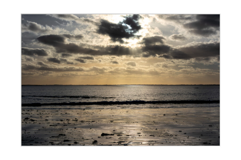 La lumière du soleil traverse des nuages spectaculaires au-dessus d'une mer calme à marée basse, se reflétant sur le sable mouillé et les rochers épars d'une plage tranquille.