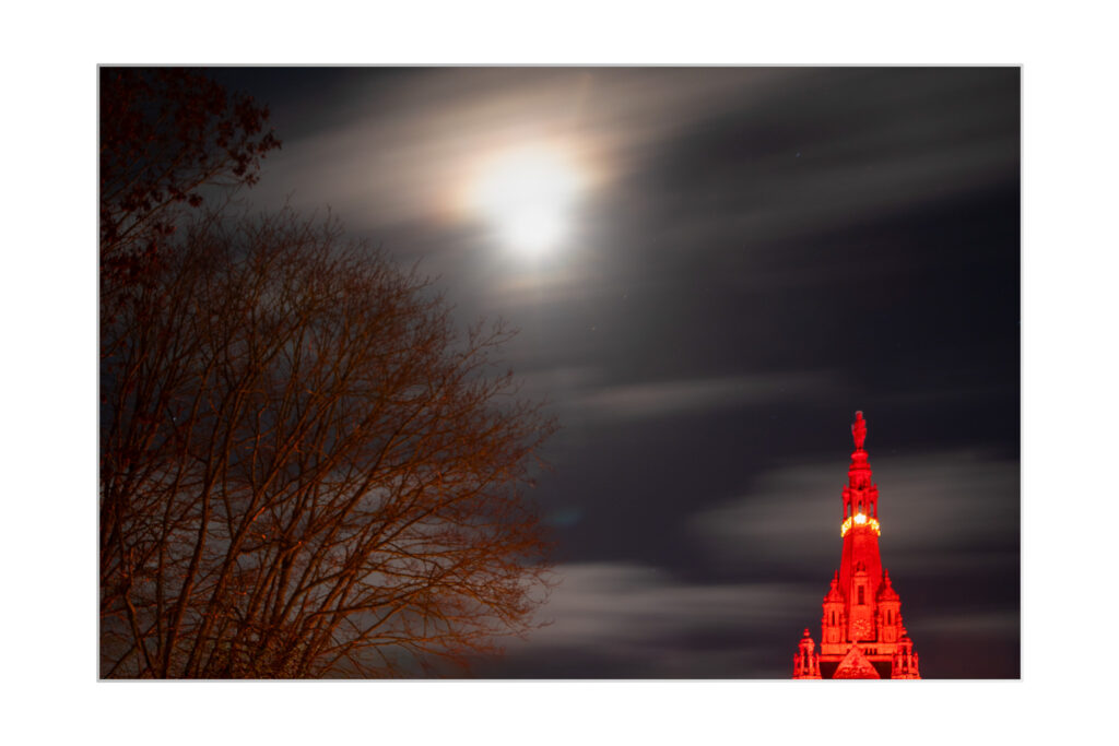 La flèche de la basilique Sainte-Anne illuminé brille en rouge la nuit sous une lune nuageuse et brillante. Des branches d'arbres dénudées encadrent le côté gauche de l'image sur fond de ciel sombre.