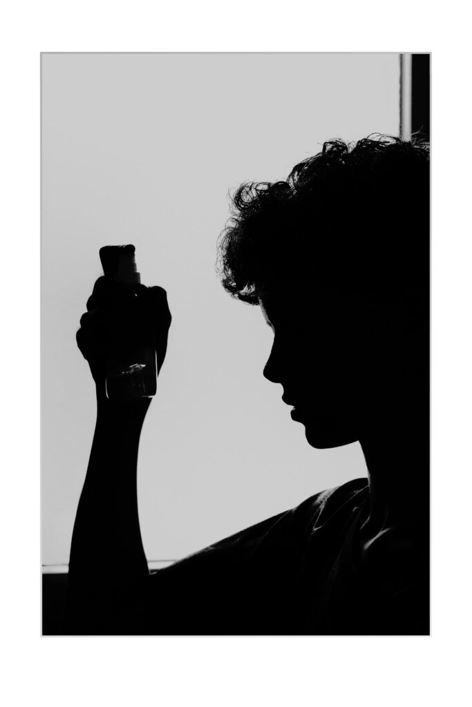 Silhouette d'une personne aux cheveux bouclés tenant un petit flacon pulvérisateur, sur un fond clair avec un cadre de fenêtre visible. Les traits du visage et les détails sont dans l'ombre.