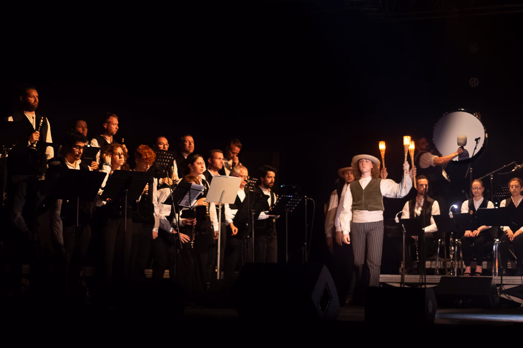 Un groupe de musiciens et de chanteurs, habillés en noir et blanc, se produit sur une scène faiblement éclairée. L'un d'entre eux tient deux torches allumées tandis que d'autres chantent et jouent d'instruments.