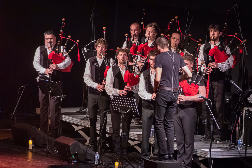 Un groupe de musiciens jouant de la cornemuse sur scène, tous vêtus de gilets de cérémonie et de chemises blanches. Un chef d'orchestre se tient devant eux et dirige la performance sous l'éclairage de la scène.
