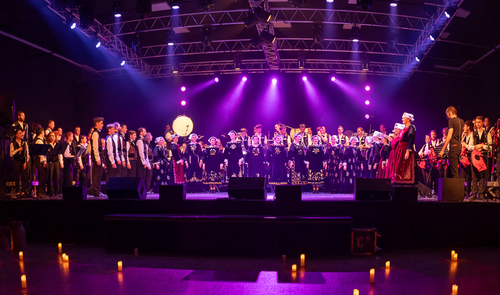 La Kevren Alré, en costumes traditionnels se tient sur une scène éclairée de lumières violettes, avec des musiciens de part et d'autre. Des bougies électriques tapissent le sol devant la scène.