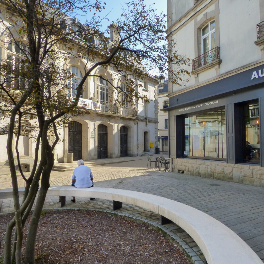 Une personne est assise seule sur un banc incurvé près d'un petit arbre, sur une place européenne calme et ensoleillée, avec en arrière-plan des bâtiments historiques et une boulangerie.