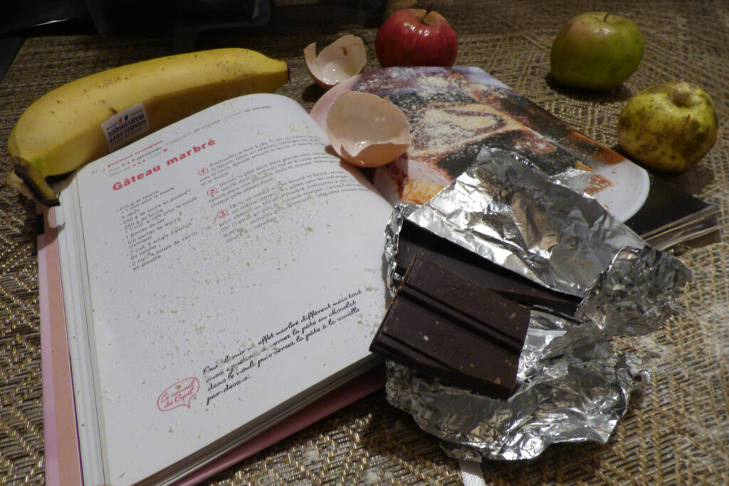Un livre de cuisine ouvert, sur lequel figure une recette de gâteau marbré, est posé sur une table aux côtés d'une banane, de deux pommes, de morceaux de chocolat emballés dans du papier d'aluminium, de coquilles d'œufs et d'un autre magazine de cuisine. La scène suggère la préparation d'une pâtisserie.