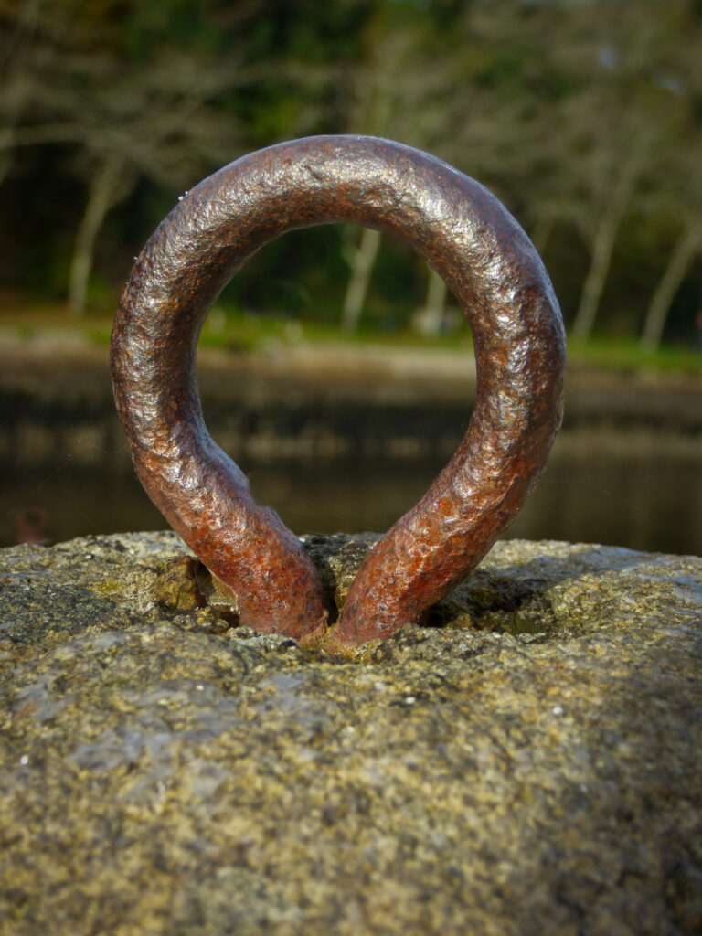 Gros plan sur une boucle métallique rouillée encastrée dans une surface de pierre brute et érodée par les intempéries, avec en arrière-plan des arbres et de l'eau flous.