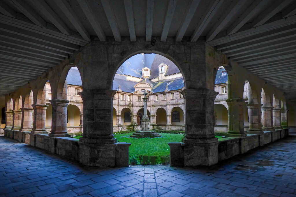 Des arcades en pierre encadrent une cour circulaire agrémentée d'une fontaine centrale et d'une pelouse verdoyante, entourée de bâtiments historiques aux fenêtres cintrées et aux toits d'ardoise, sous un ciel couvert.