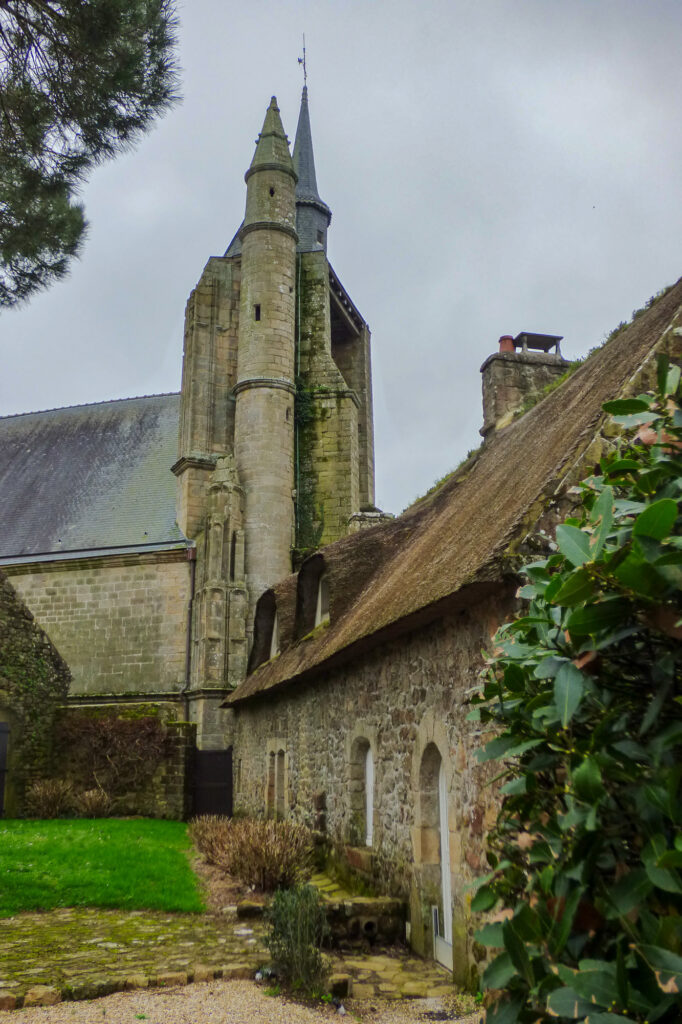 Une église en pierre, surmontée d'un haut clocher pointu, se dresse à côté d'un gîte rustique en pierre au toit de chaume, au milieu d'un écrin de verdure sous un ciel nuageux.