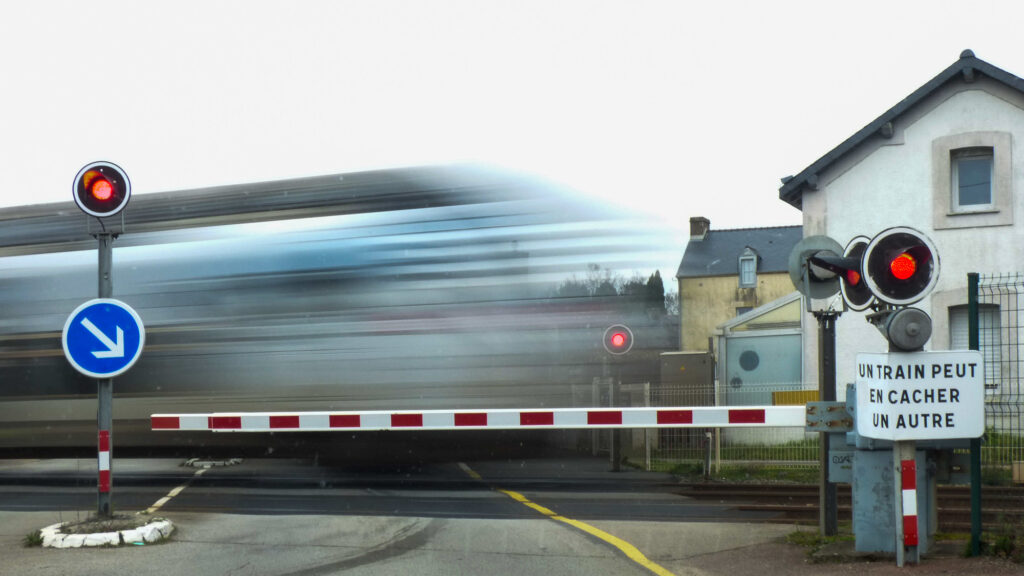 Un train filant à toute allure passe à toute vitesse devant une barrière abaissée à un passage à niveau équipé de feux rouges. Un panneau en français avertit : « UN TRAIN PEUT EN CACHER UN AUTRE ». On aperçoit des maisons à l'arrière-plan.