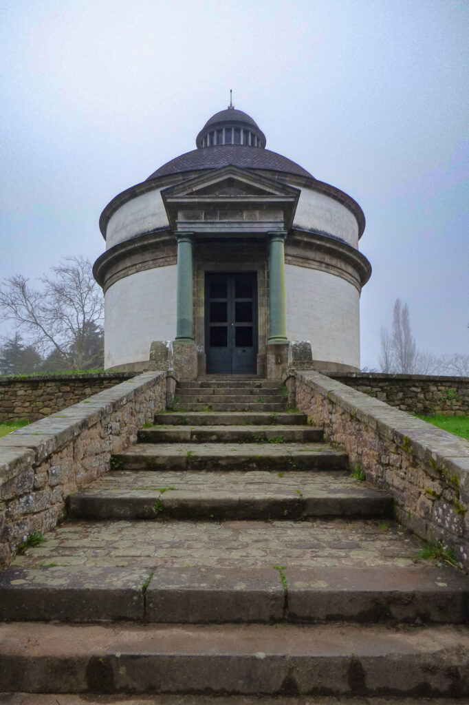 Un escalier en pierre mène à un petit édifice néoclassique de forme circulaire, doté d'un toit en dôme et de colonnes à l'entrée, se découpant sur un ciel nuageux et couvert, avec des arbres dénudés en arrière-plan.