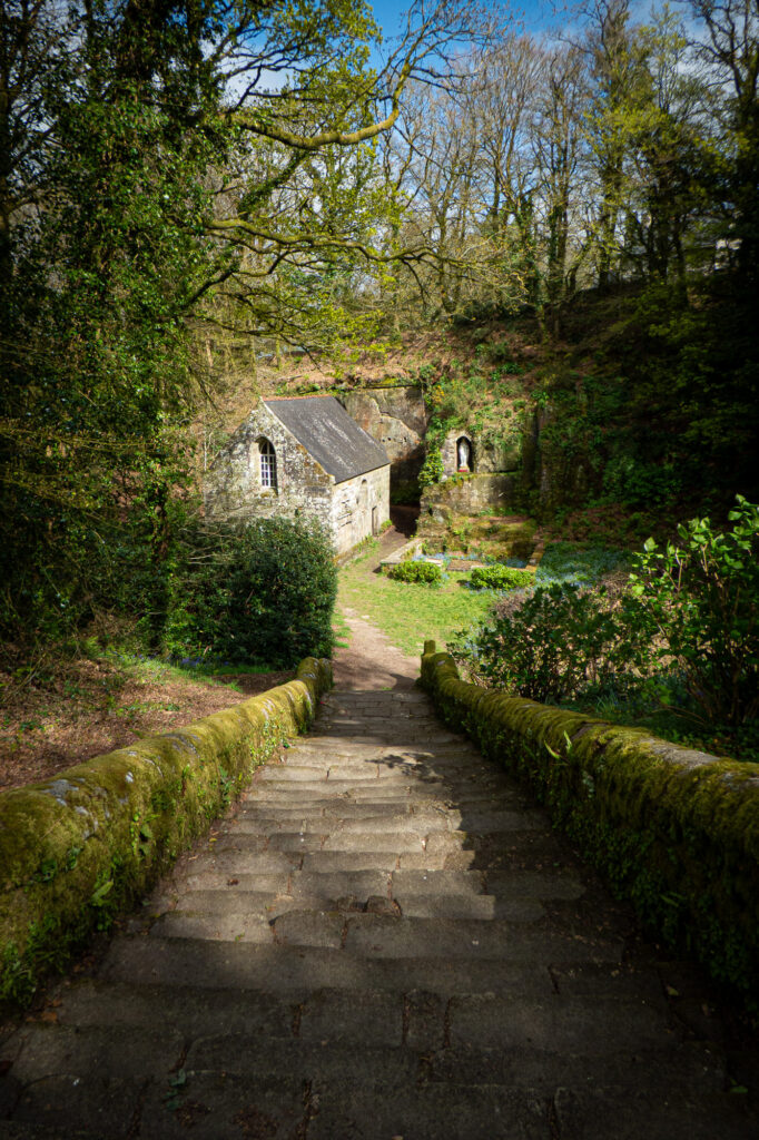 Un sentier de pierre recouvert de mousse descend vers une petite chapelle en pierre nichée au cœur d'une forêt verdoyante et luxuriante, peuplée de grands arbres au feuillage dense, sous un ciel partiellement nuageux.