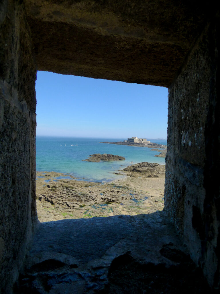 Une fenêtre en pierre encadre la vue sur une plage rocheuse aux eaux d'un bleu limpide, avec au loin une vieille forteresse sur une île, sous un ciel d'un bleu éclatant.