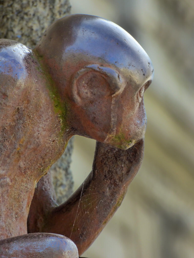 Gros plan sur une sculpture en bronze patinée représentant un singe assis, la main portée à la bouche, l'air pensif. De la mousse et des toiles d'araignée sont visibles sur la statue, et l'arrière-plan est flou.