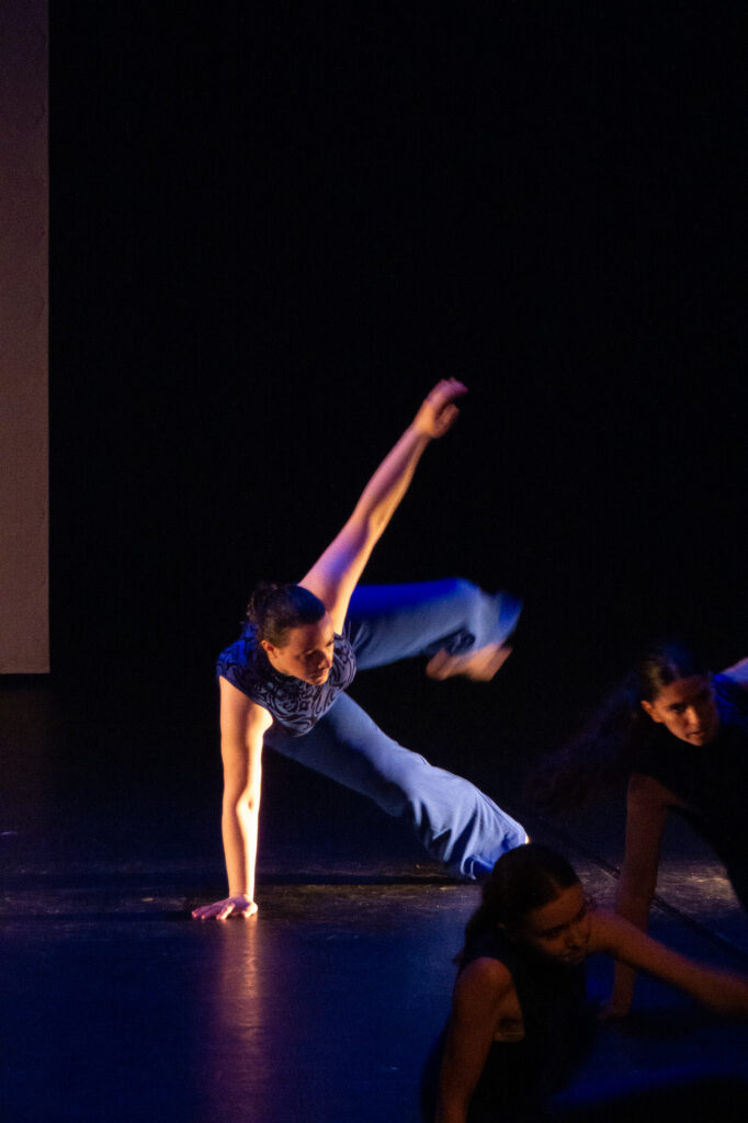 Une danseuse en pantalon bleu se tient en équilibre sur une main, l'autre bras et la jambe tendus, sur une scène sombre, tandis que deux autres danseurs sont partiellement visibles au premier plan.