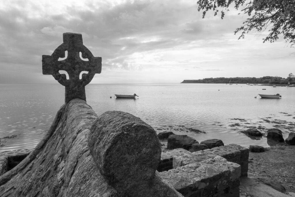 Une croix celtique en pierre se dresse sur un mur en pente, près d'un rivage rocheux, surplombant des eaux calmes où flottent quelques petits bateaux, avec au loin une lisière de forêt sous un ciel nuageux. Photographie en noir et blanc.