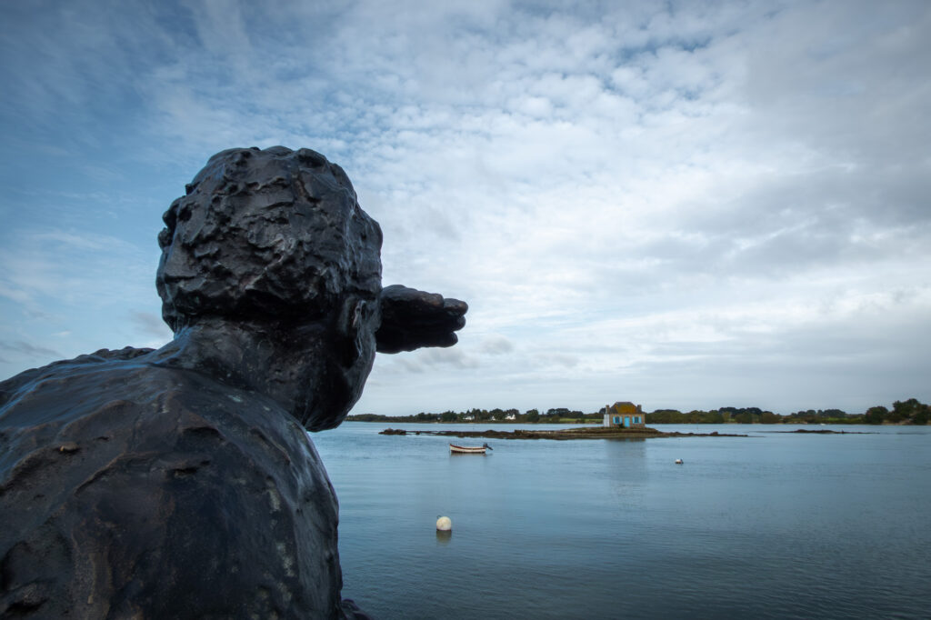 Une statue en bronze représentant une personne qui se protège les yeux de la lumière et contemple, au-delà d'un plan d'eau calme, une petite île sur laquelle se dresse une maison bleue, sous un ciel nuageux. Un bateau flotte sur l'eau à proximité.