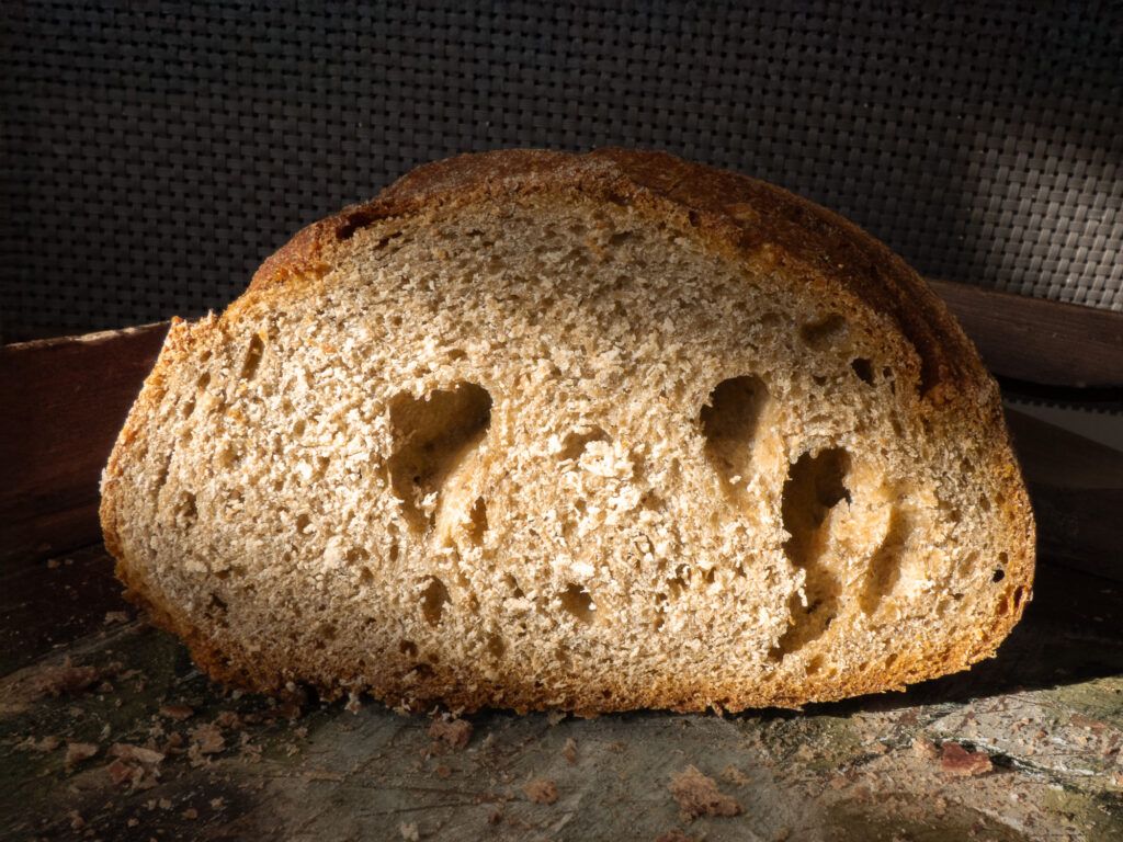 Gros plan sur une tranche de pain de campagne brun à la croûte croustillante, mettant en valeur sa texture aérée et ses alvéoles, posée sur une surface en bois parsemée de miettes.