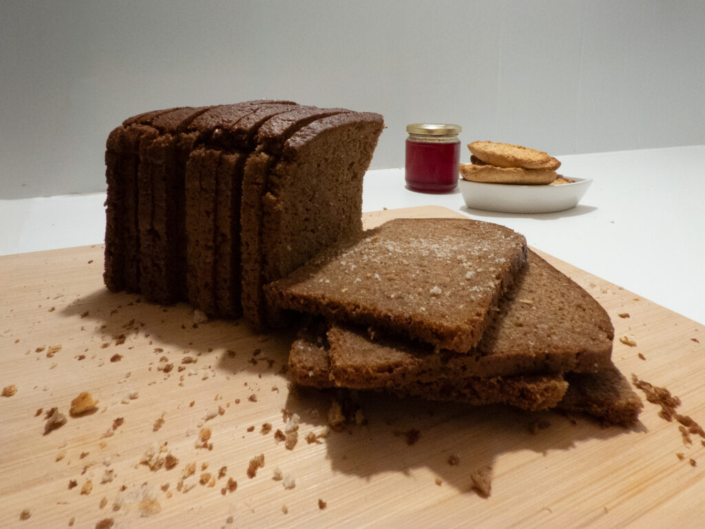 Des tranches de pain complet posées sur une planche en bois parsemée de miettes, un pot de confiture rouge et, à l'arrière-plan, une petite assiette garnie de biscuits ronds.