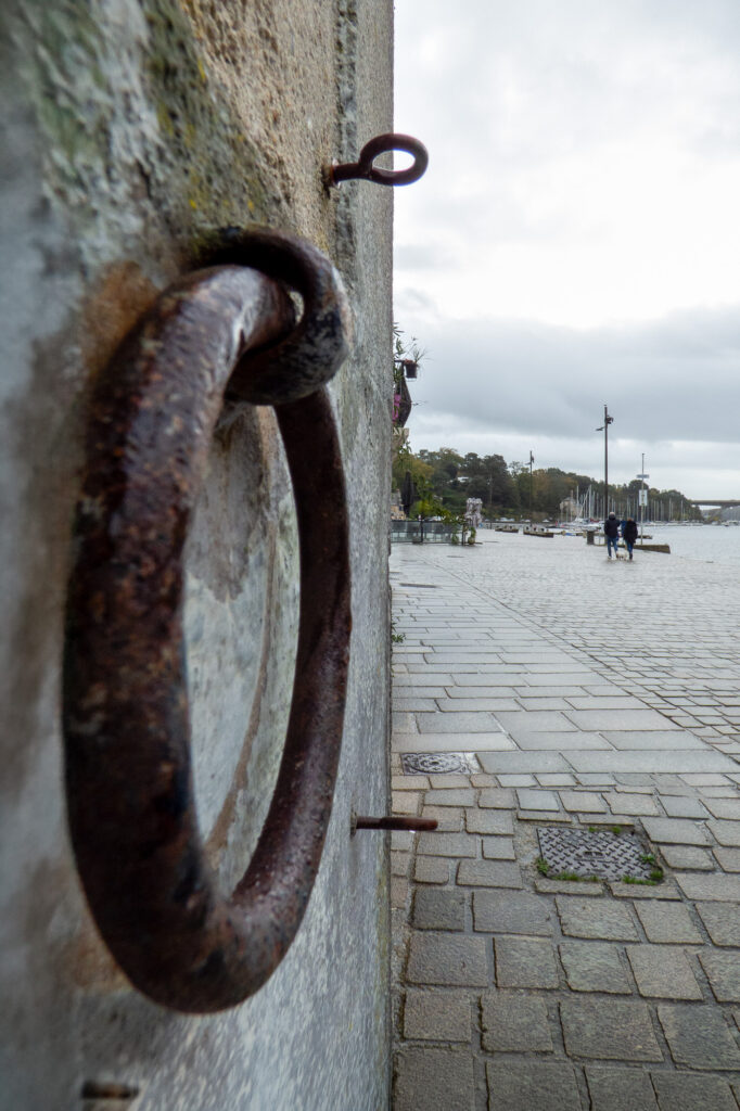 Gros plan sur un anneau métallique rouillé fixé à un mur de pierre au bord d'un quai pavé, avec au loin des passants marchant près de bateaux sous un ciel nuageux.
