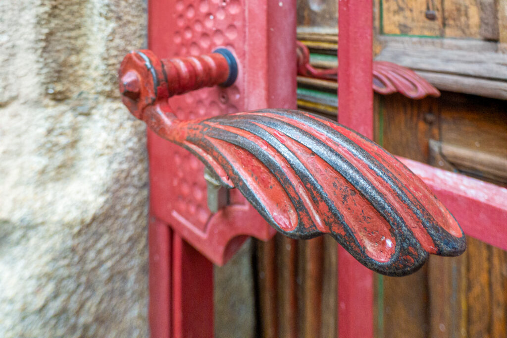 Gros plan sur une poignée de porte en métal rouge et noir, richement décorée et ornée d'un motif en forme d'ailes, fixée à une porte rouge, avec en arrière-plan un mur en pierre et une surface en bois.