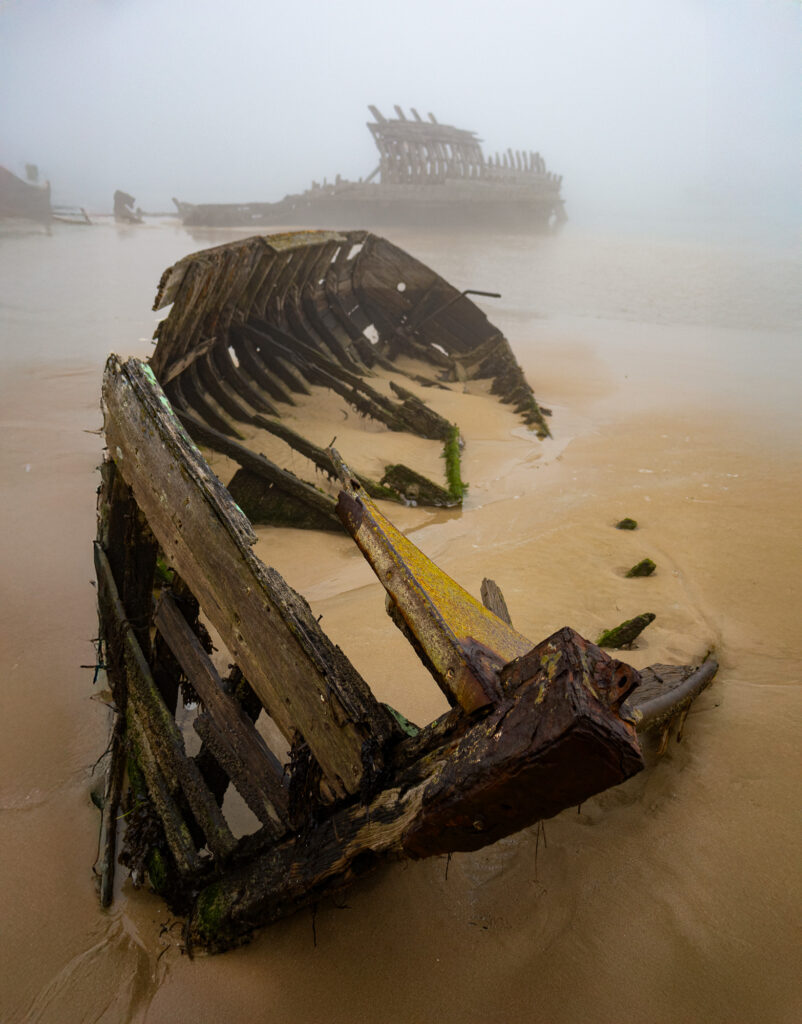 La photo montre les vestiges squelettiques et érodés d'épaves en bois éparpillées le long d'une plage de sable enveloppée de brume. Le brouillard voile l'arrière-plan, créant une scène mystérieuse et pleine d'ambiance.