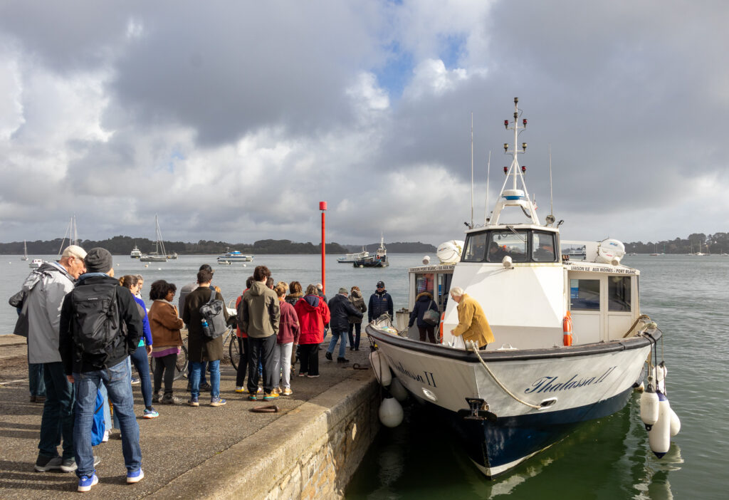 Un groupe de personnes se tient sur un quai, près d'un bateau blanc baptisé « Ithalassa II », s'apprêtant à monter à bord. Le ciel est partiellement nuageux, et plusieurs bateaux sont amarrés sur l'eau, à l'arrière-plan.