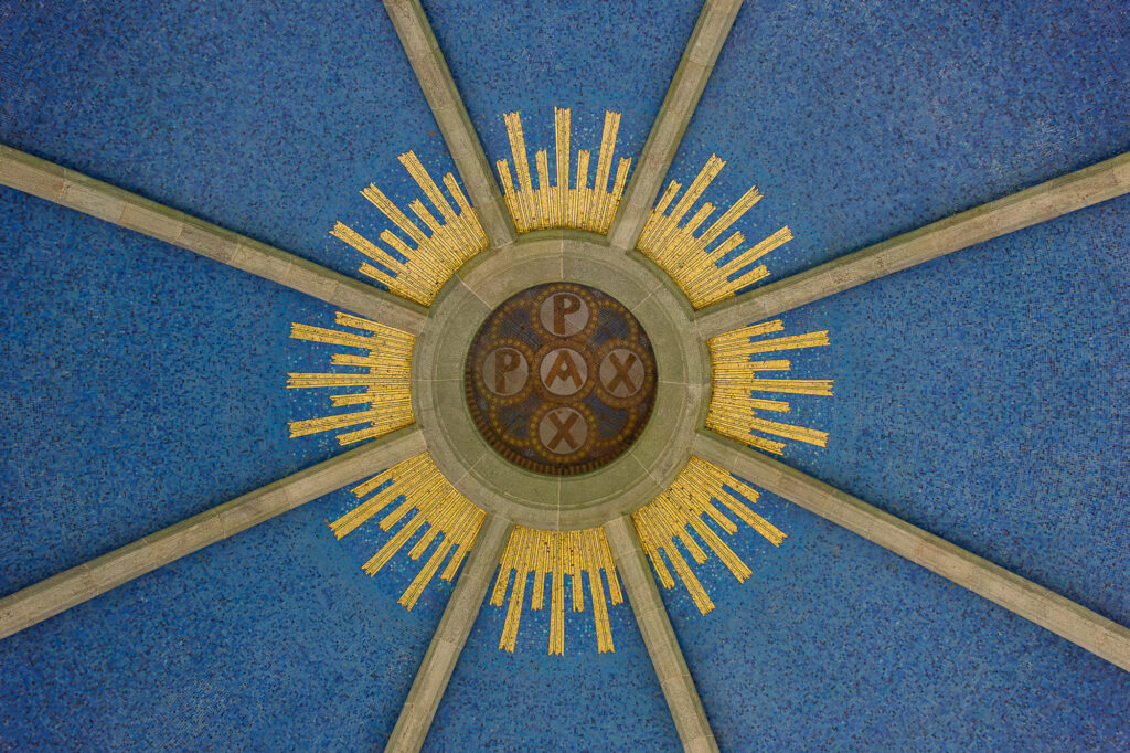 Le motif circulaire du plafond met en avant le mot « PAX » au centre, entouré de rayons dorés sur un fond bleu texturé, le tout divisé par des poutres de soutien grises rayonnant vers l'extérieur.