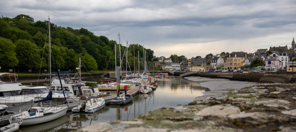 Un petit port où des voiliers et des yachts sont amarrés au bord d'une eau calme, bordé d'arbres et de bâtiments pittoresques sous un ciel nuageux, avec un mur de pierre au premier plan.