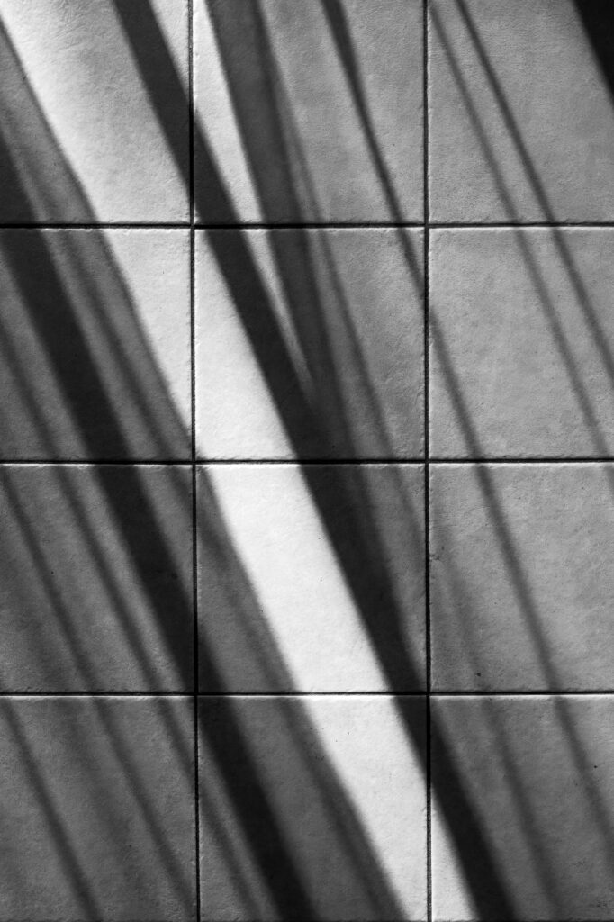 Une grille de carreaux carrés sur lesquels se projettent des ombres en diagonale. Ces ombres créent un motif alternant zones claires et zones sombres sur la surface carrelée, par ailleurs lisse et de couleur claire.