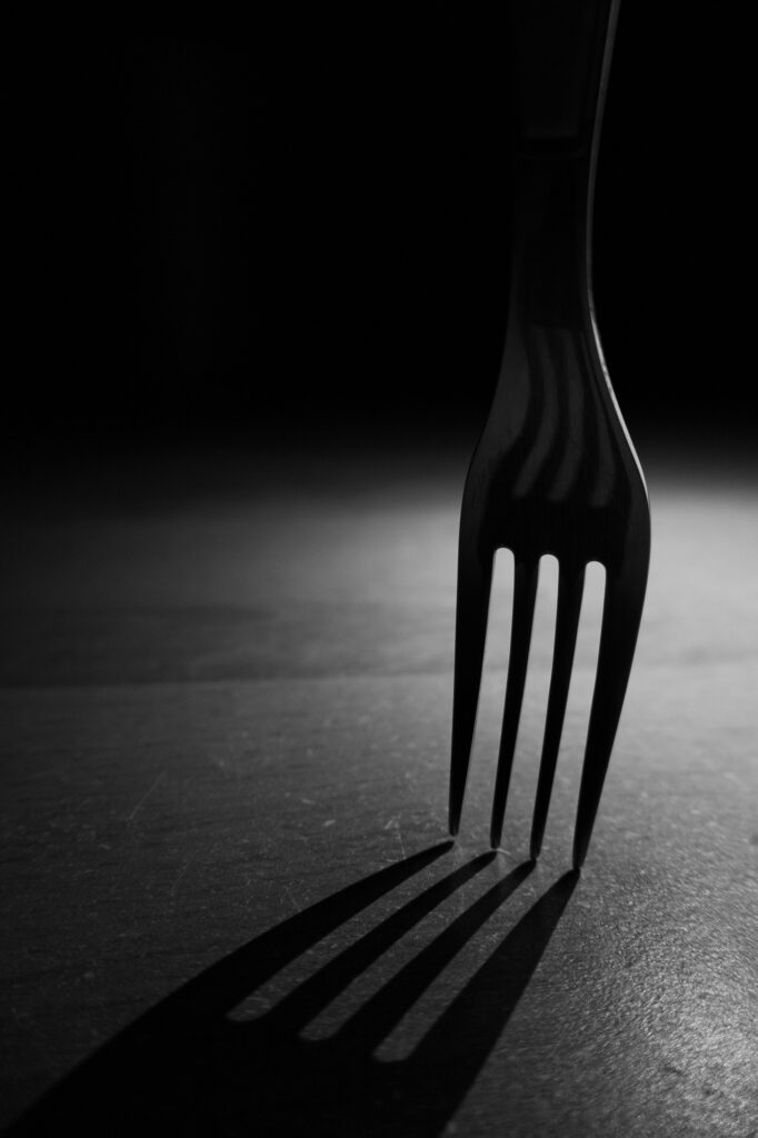 Une fourchette posée à la verticale sur une surface, éclairée de manière spectaculaire de sorte que ses dents projettent de longues ombres sombres sur un fond sombre.