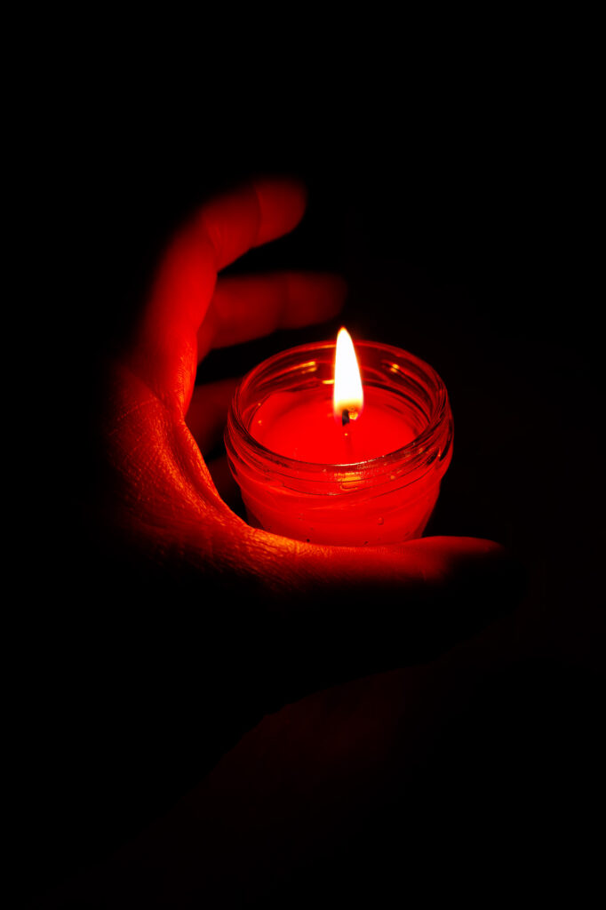 Une main tient un petit bocal en verre dans lequel brûle une bougie rouge, qui diffuse une lueur chaleureuse dans l'obscurité environnante.