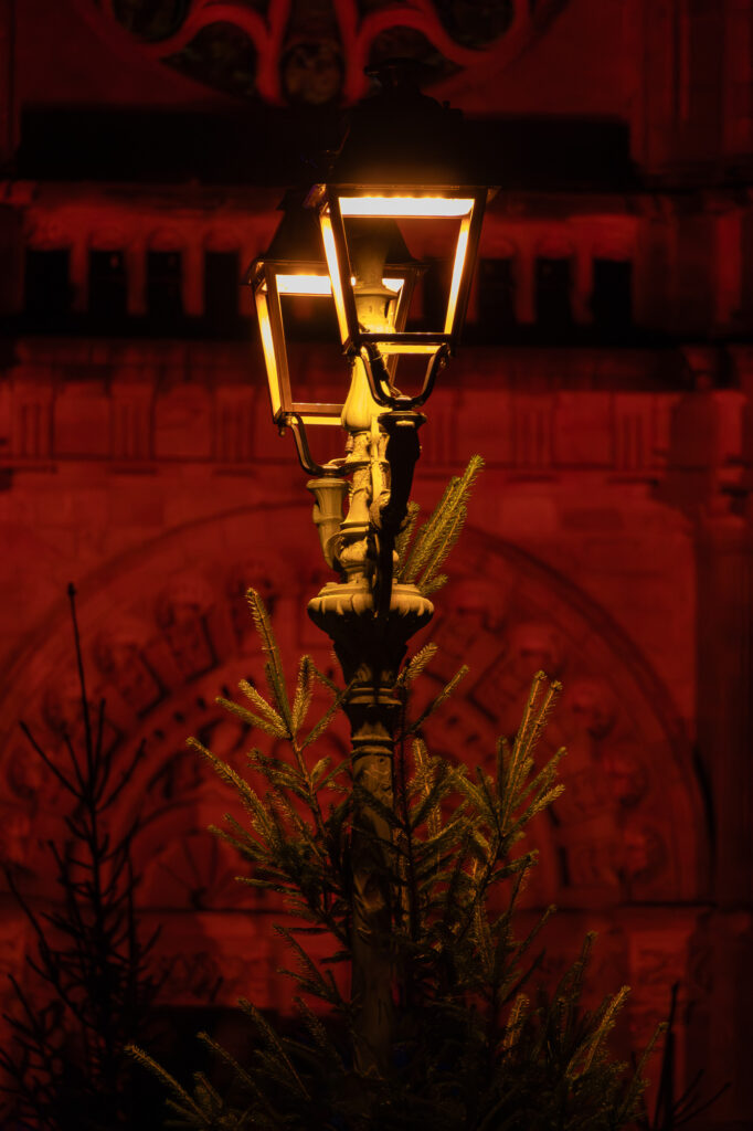 Un lampadaire ancien diffuse une lumière jaune et chaleureuse la nuit, entouré de branches de pin. En arrière-plan, on aperçoit un bâtiment en pierre richement décoré, baigné d'une lumière rouge foncé.