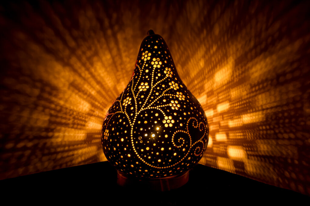 Une lampe décorative en calebasse, ornée de motifs découpés très travaillés, diffuse une lumière chaleureuse, projetant des jeux de lumière tourbillonnants et des ombres géométriques sur les murs environnants.